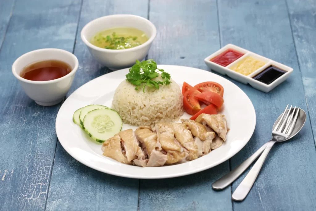  Hainanese chicken rice served with tomatoes, cucumbers, garnish, sauces, and soups laid on a grey wooden table

