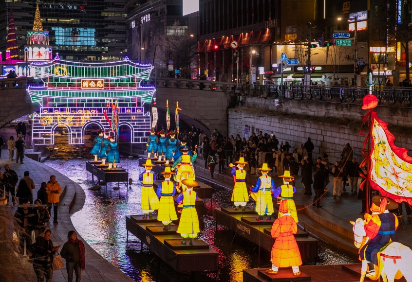 Illuminated lantern figures depicting traditional Korean characters displayed along Cheonggyecheon Stream in Seoul at night during a lantern festival.